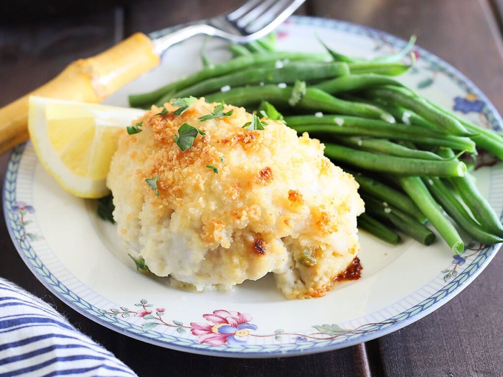 Stuffed catfish fillet on a floral plate with green beans and a lemon wedge. Glass of white wine and a skillet with additional catfish fillets in the background.