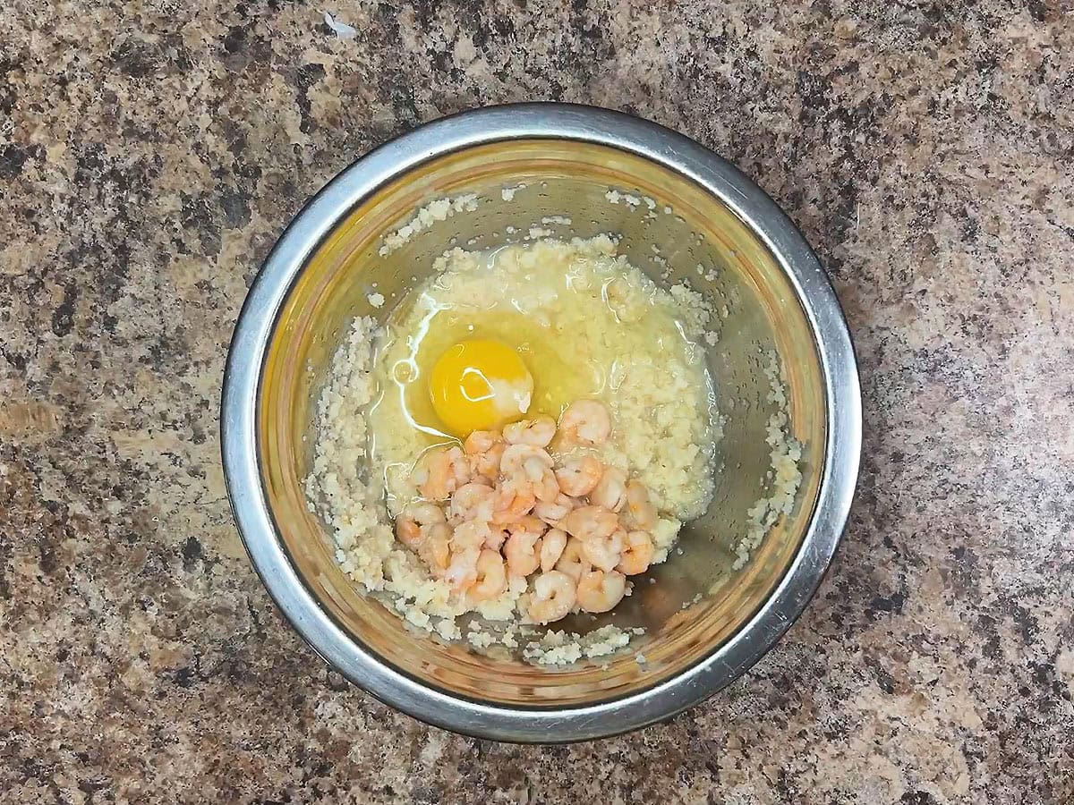 Mashed cornbread, a whole egg, and small shrimp in a small mixing bowl.