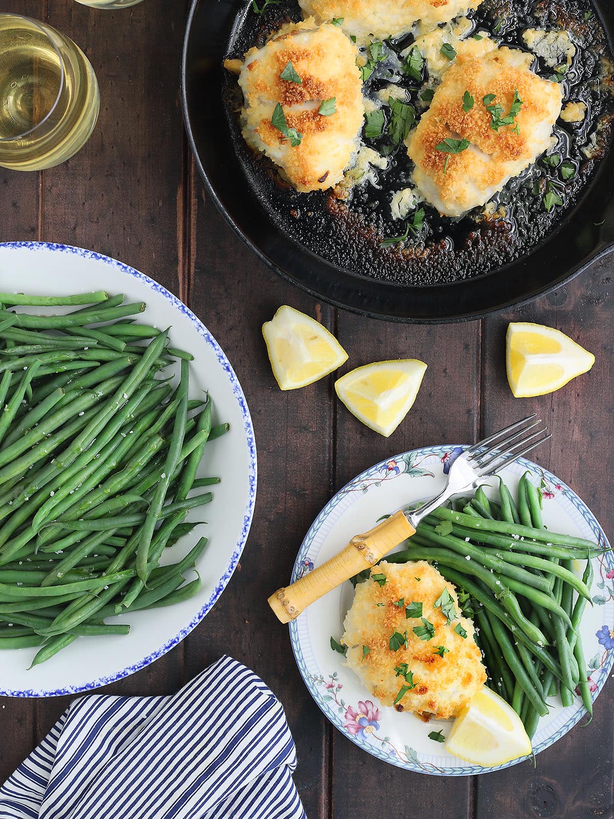 Overhead shot of a stuffed catfish fillet on a floral plate with green beans and a lemon wedge. Glass of white wine, a skillet with additional catfish fillets, and a bowl of green beans on the side.