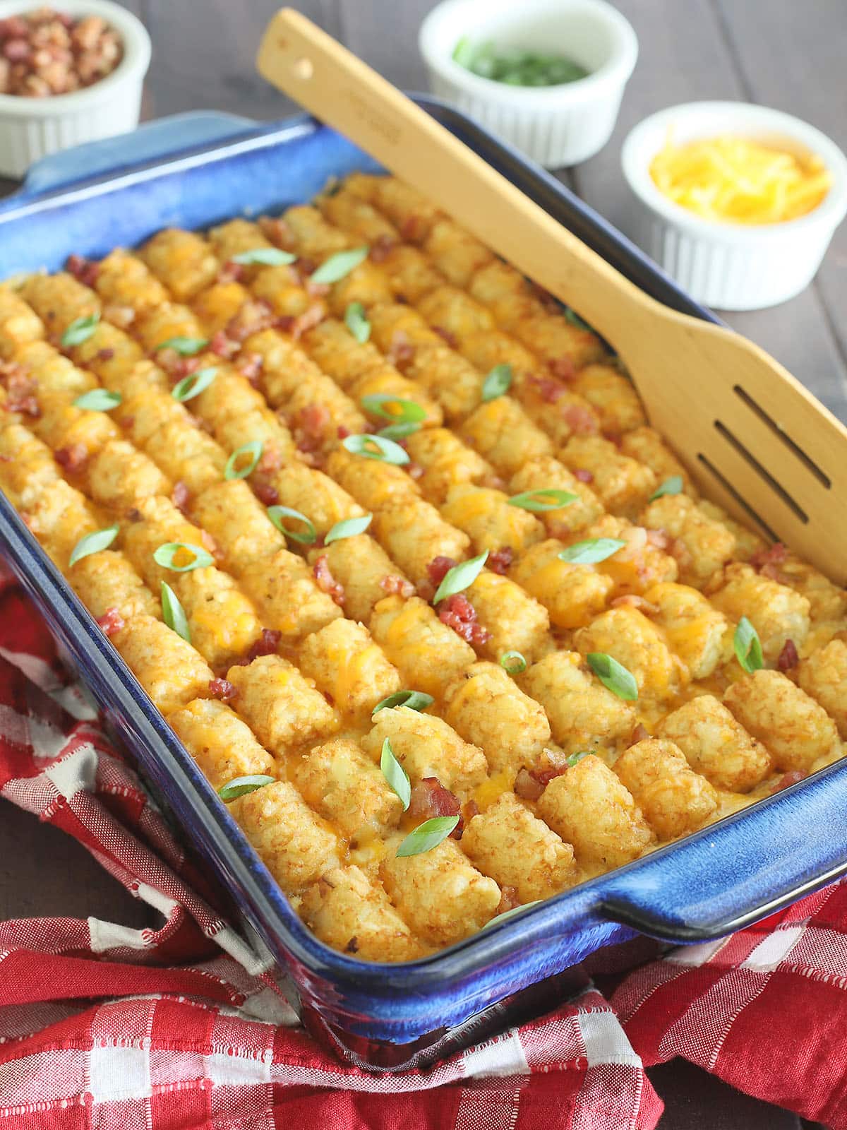 Bacon chicken ranch tater tot casserole garnished with sliced green onions. A wooden spatula is tucked into the side of the casserole. Small dishes of additional green onions, bacon bits, and shredded cheese in the background.
