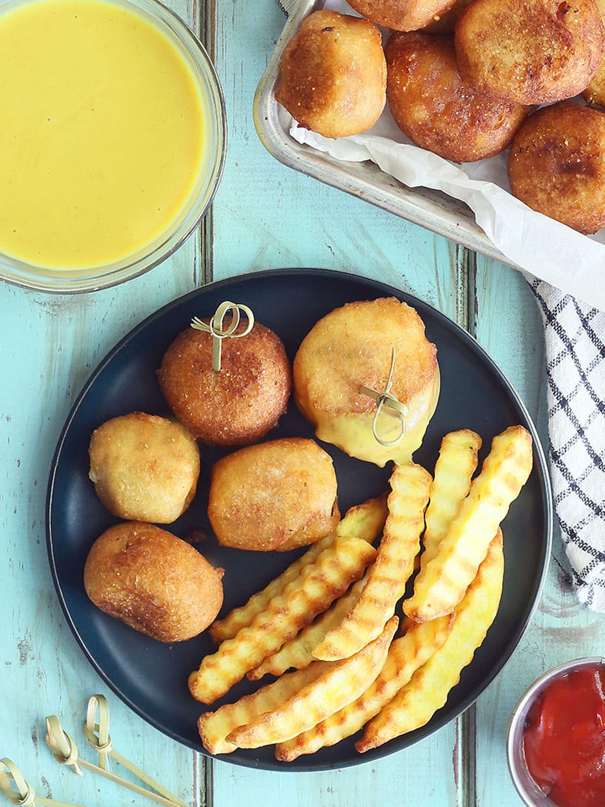 Five mini corndog bites on a plate with crinkle cut French fries. Half of one corndog bite is coated in honey mustard sauce. A dish of honey mustard sauce, ketchup, toothpicks, and a platter of corndog bites are to the side of the plate.