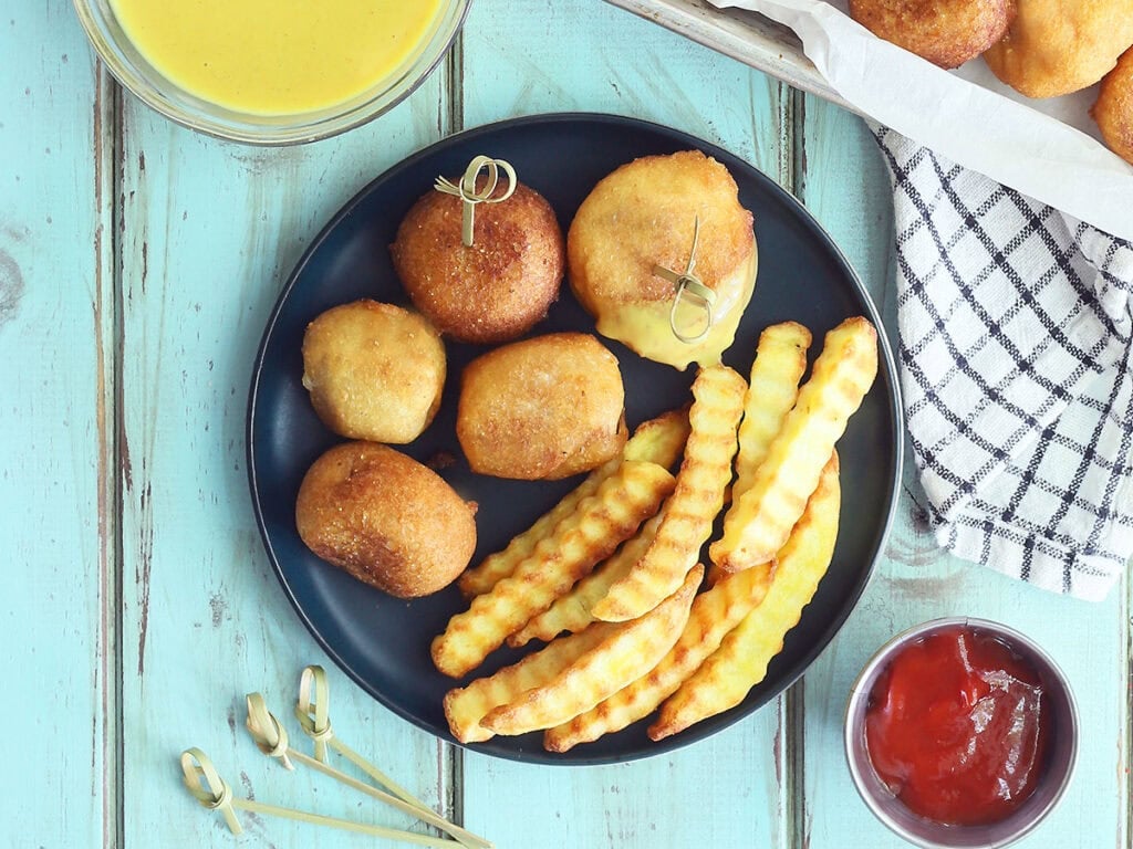Five mini corndog bites on a plate with crinkle cut French fries. Half of one corndog bite is coated in honey mustard sauce. A dish of honey mustard sauce, ketchup and toothpicks are to the side of the plate.
