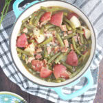 Green beans with ham and potatoes in a Dutch oven with a napkin, wooden spoon, plates with forks, and a few sprigs of parsley to the side.