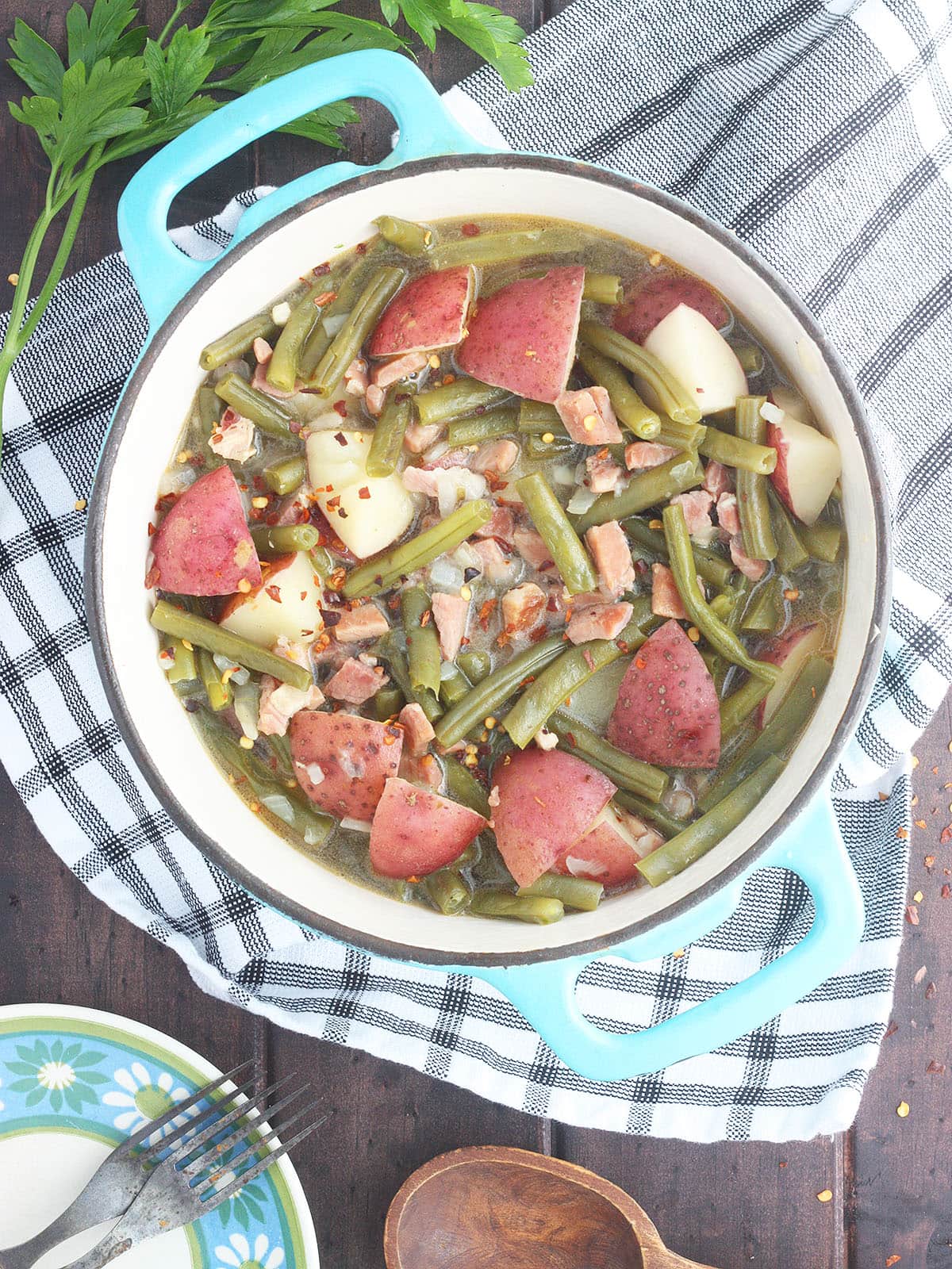 Green beans with ham and potatoes in a Dutch oven with a napkin, wooden spoon, plates with forks, and a few sprigs of parsley to the side.