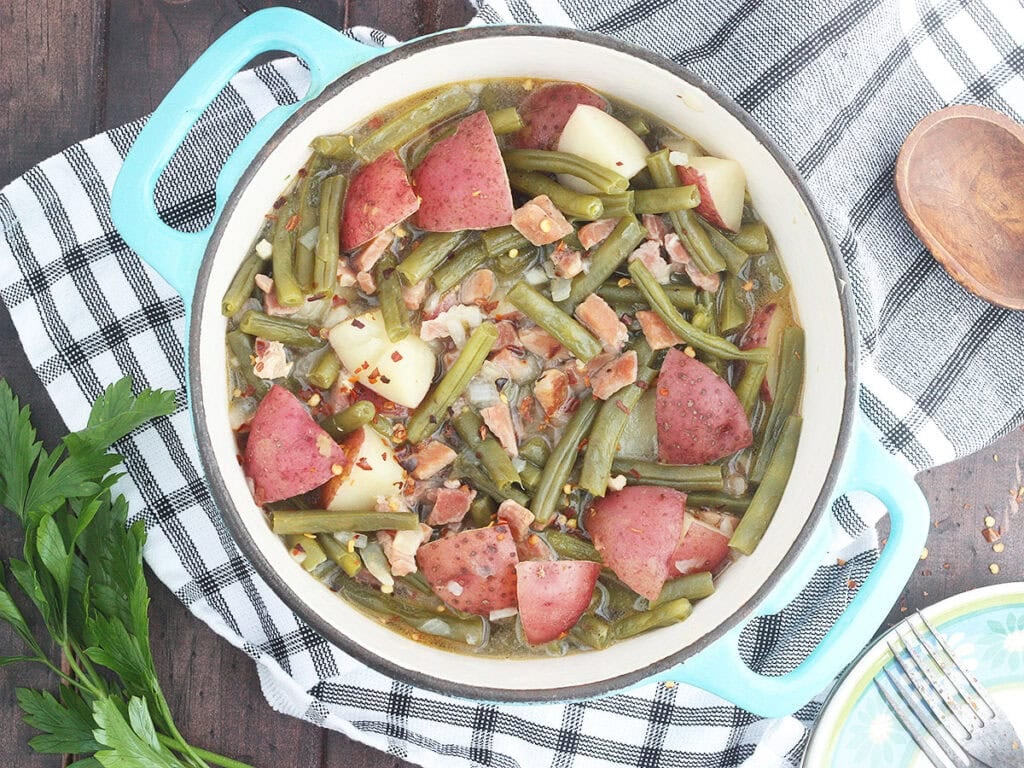 Green beans with ham and potatoes in a Dutch oven with a napkin, wooden spoon, and a few sprigs of parsley to the side.