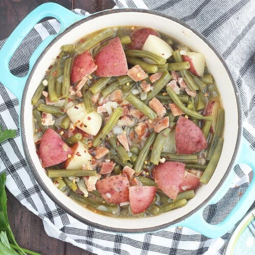 Green beans with ham and potatoes in a Dutch oven with a napkin, wooden spoon, and a few sprigs of parsley to the side.
