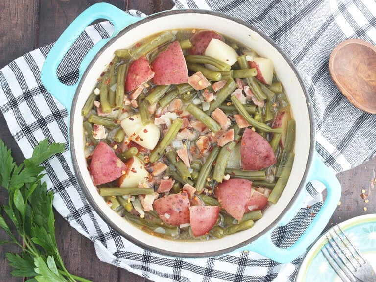 Green beans with ham and potatoes in a Dutch oven with a napkin, wooden spoon, and a few sprigs of parsley to the side.