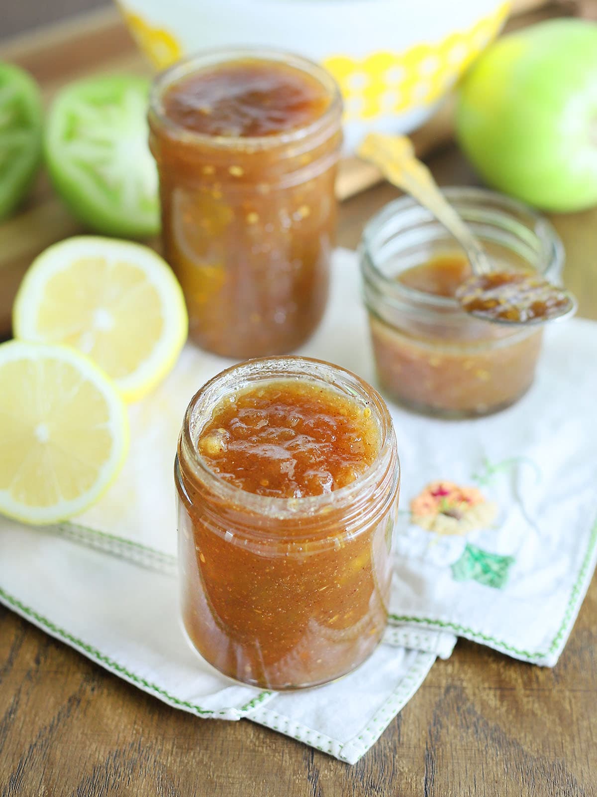 Jars of green tomato jam surrounded by sliced lemons and whole green tomatoes.