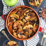 Baked apples garnished with raisins and pecans in a round clay serving dish. Smaller dishes of baked apples, a whole Granny Smith apple, a dish of brown sugar, pecans, and a measuring cup of raisins to the side.