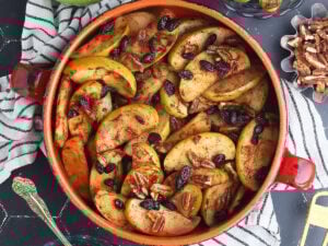 Baked apples garnished with raisins axnd pecans in a round clay serving dish. A small dish of pecans to the side.