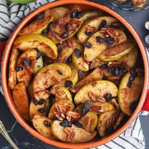 Baked apples garnished with raisins axnd pecans in a round clay serving dish. A small dish of pecans to the side.