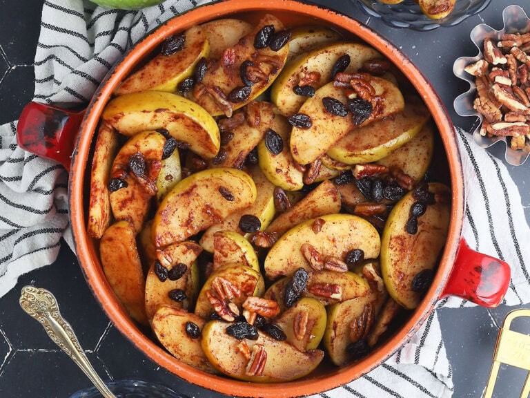 Baked apples garnished with raisins and pecans in a round clay serving dish. A small dish of pecans to the side.