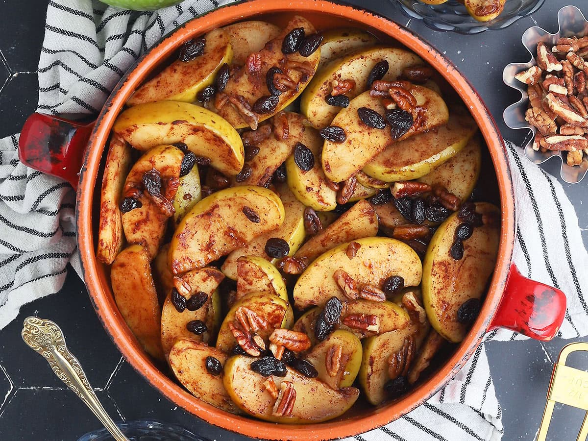 Baked apples garnished with raisins axnd pecans in a round clay serving dish. A small dish of pecans to the side.