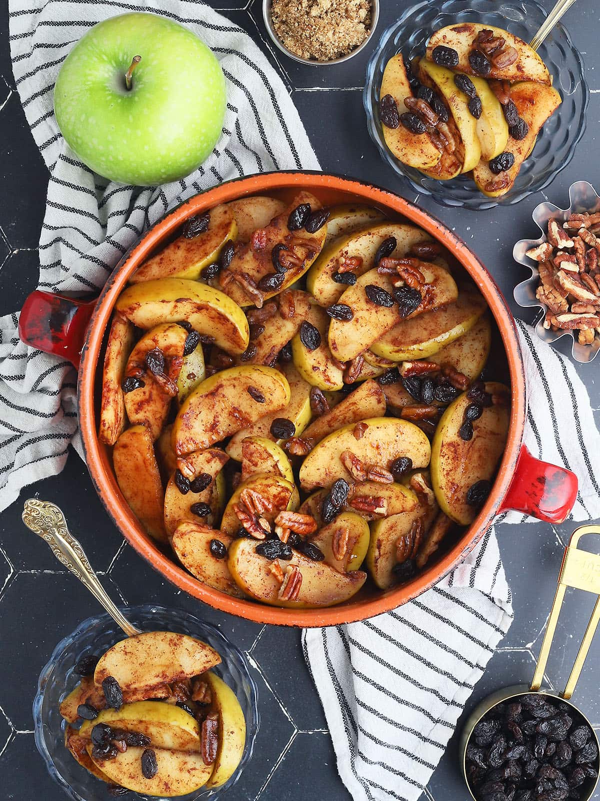 Baked apples garnished with raisins and pecans in a round clay serving dish. Smaller dishes of baked apples, a whole Granny Smith apple, a dish of brown sugar, pecans, and a measuring cup of raisins to the side.