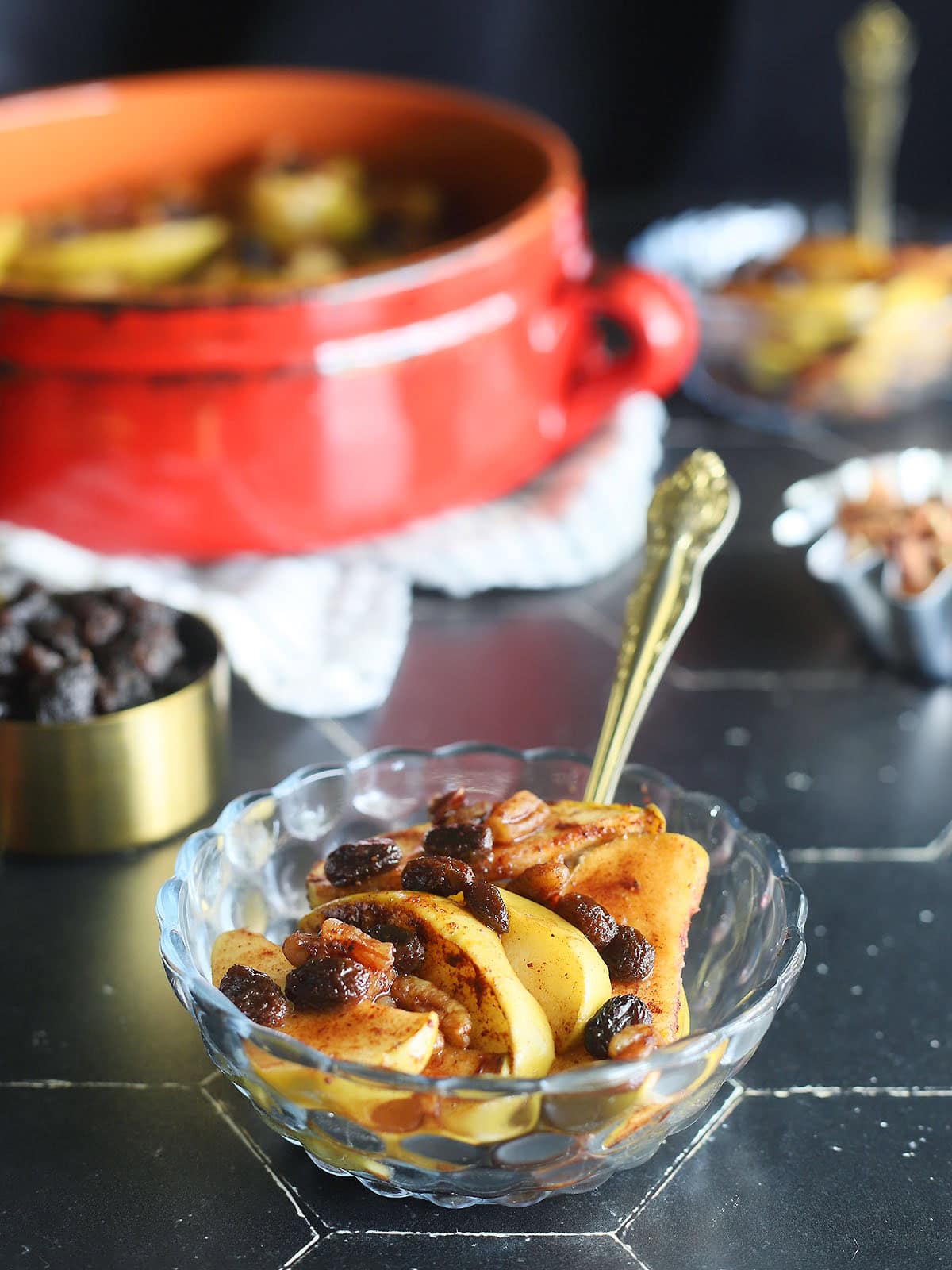 Small dish of baked apples garnished with raisins and pecans. Serving bowl of apples, measuring cup of raisins, a second dish of apples and a dish of pecans in the background.