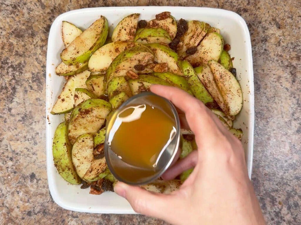 Pouring apple cider over the apples in a square baking dish.