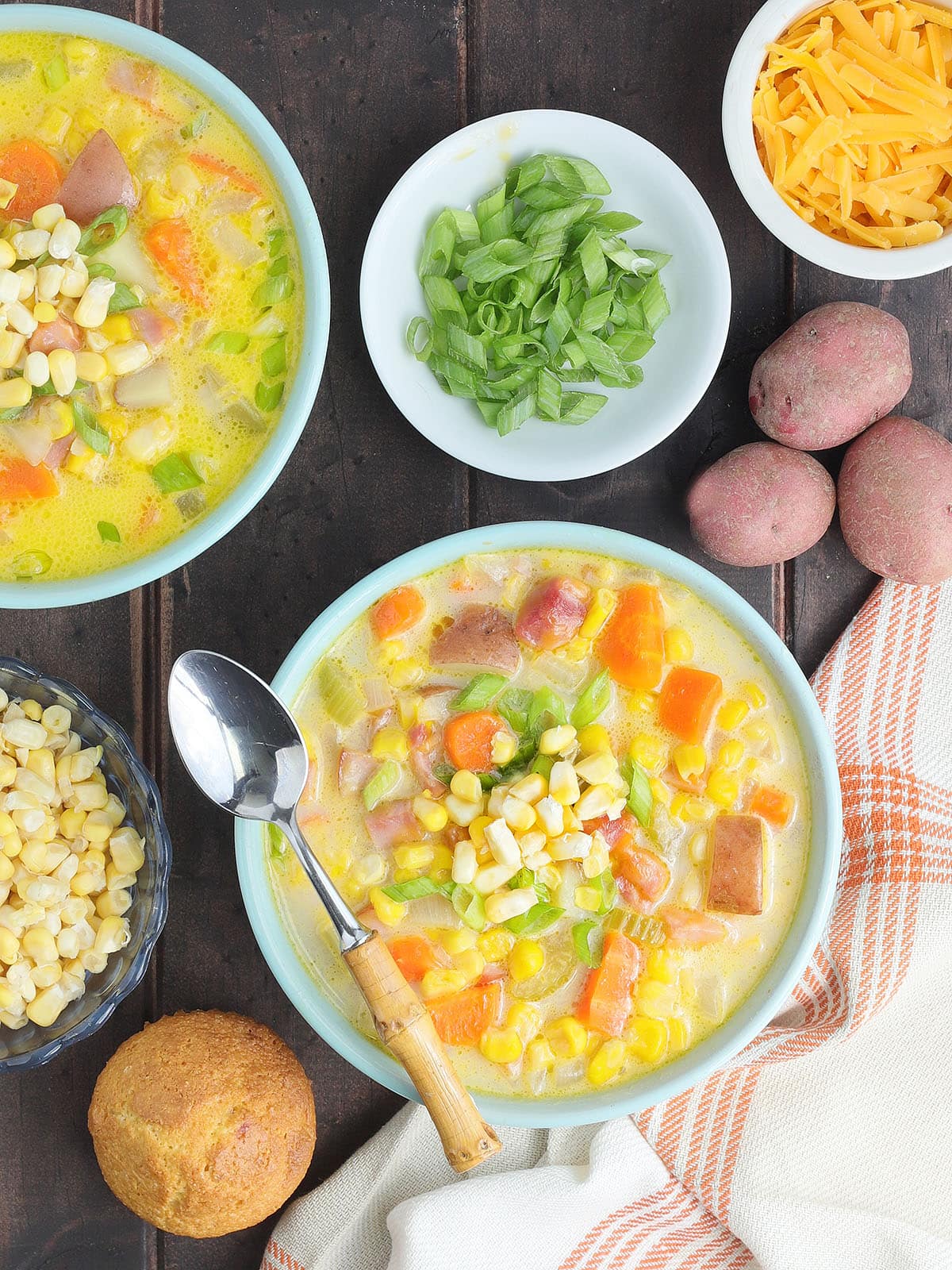 Two bowls of potato corn chowder with dishes of green onions, corn and cheese to the side.