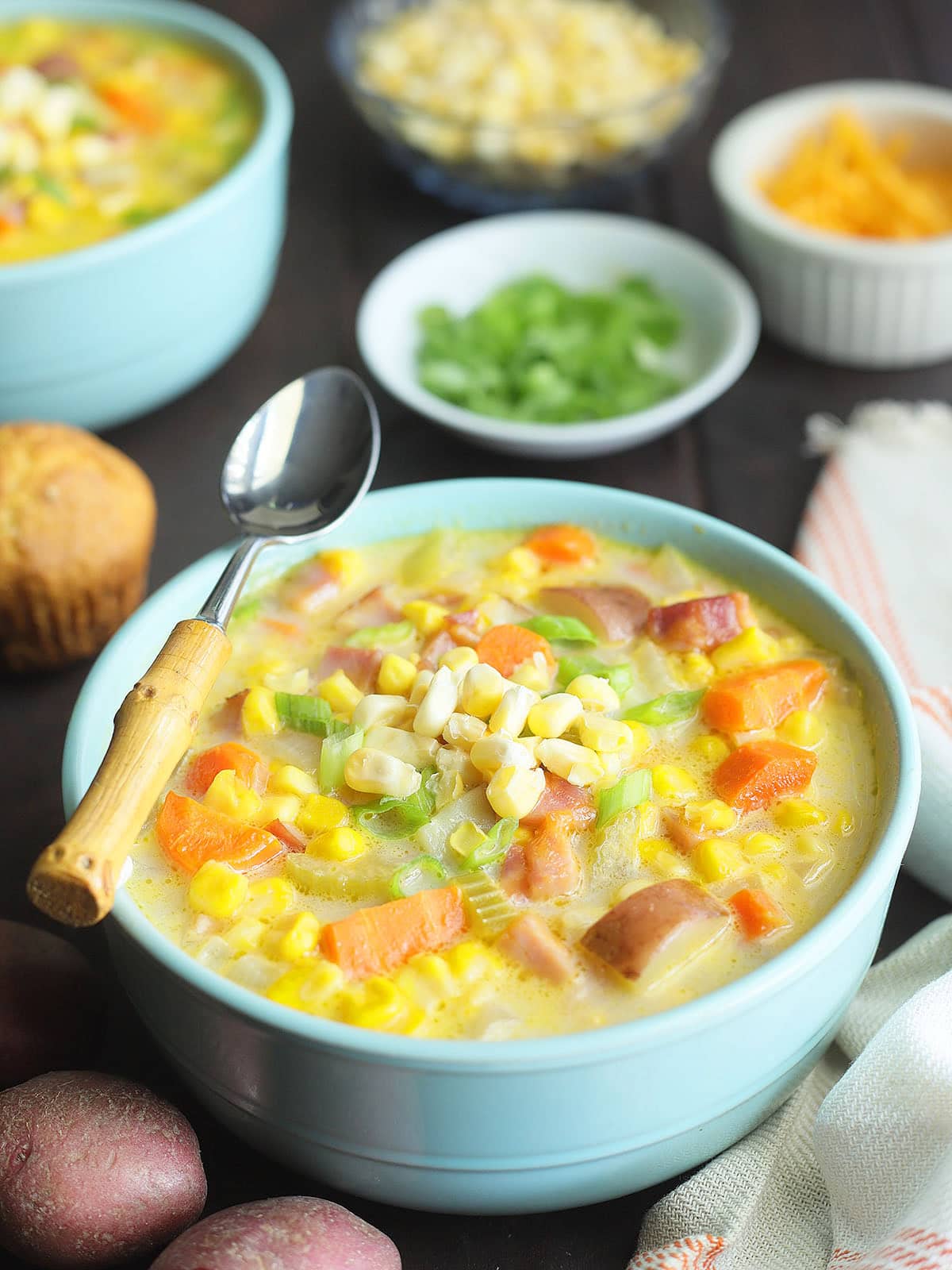 Two bowls of potato corn chowder with dishes of green onions, corn and cheese to the side.