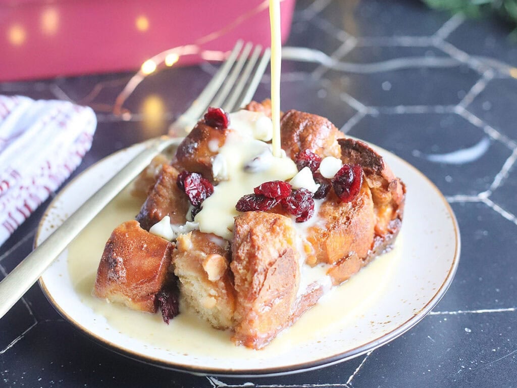 Pouring rum creme anglaise over the top of a slice of white chocolate cranberry bread pudding.