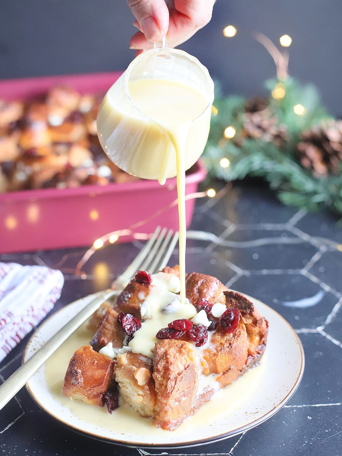 Pouring rum creme anglaise over the top of a slice of white chocolate cranberry bread pudding.