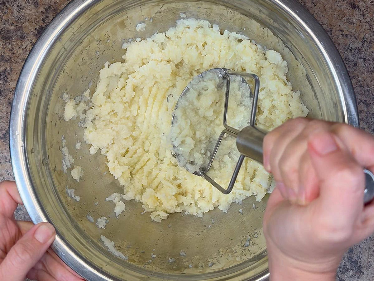 Using a potato masher to mash steamed cauliflower florets in a large mixing bowl.