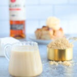 Small glass pitcher of rum sauce with a cup of brown sugar, slice of bread pudding and a bottle of rum in the background.