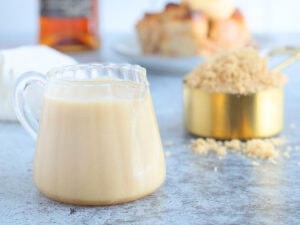 Small glass pitcher of rum sauce with a cup of brown sugar, slice of bread pudding and a bottle of rum in the background.