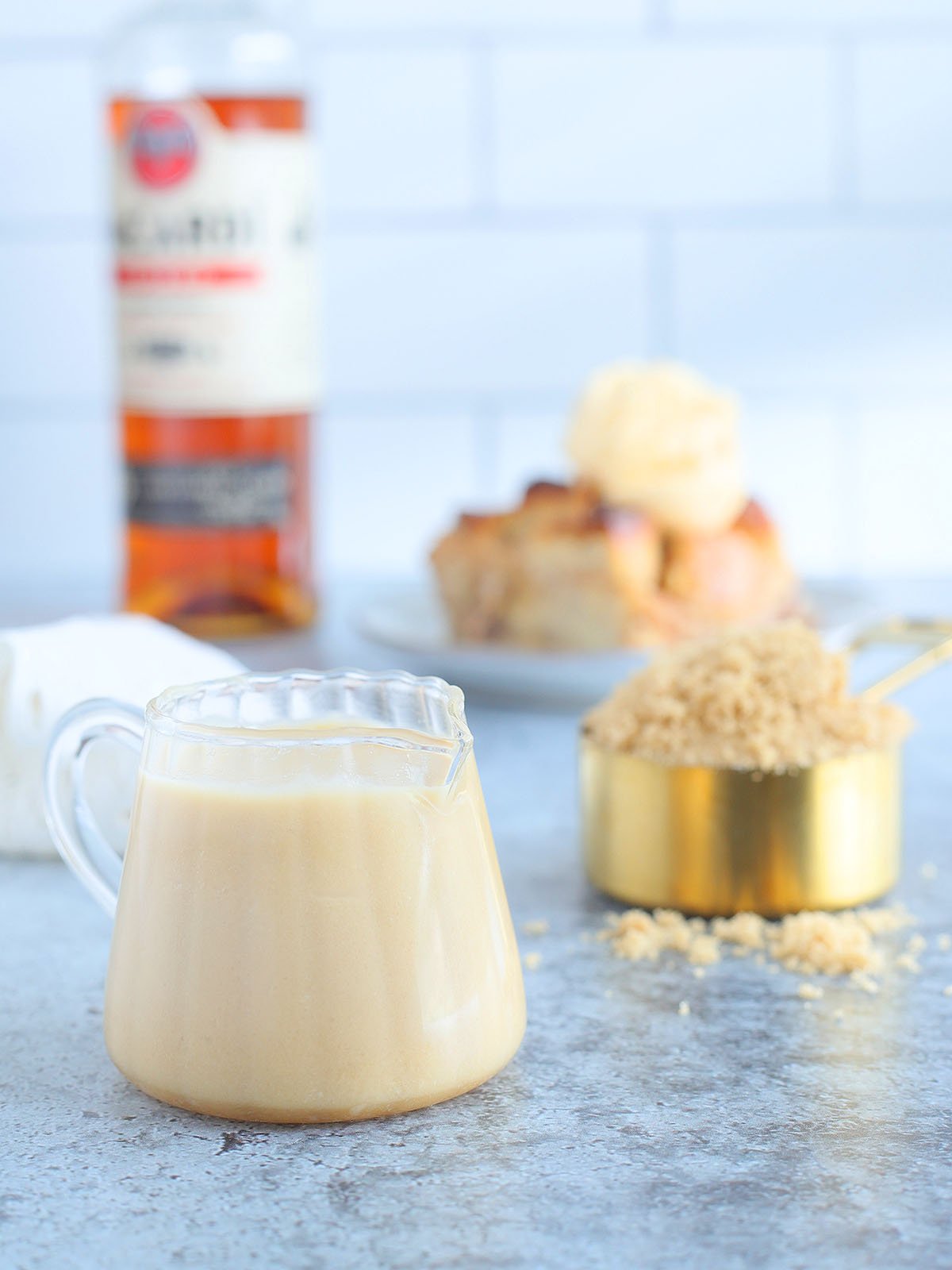 Small glass pitcher of rum sauce with a cup of brown sugar, slice of bread pudding and a bottle of rum in the background.
