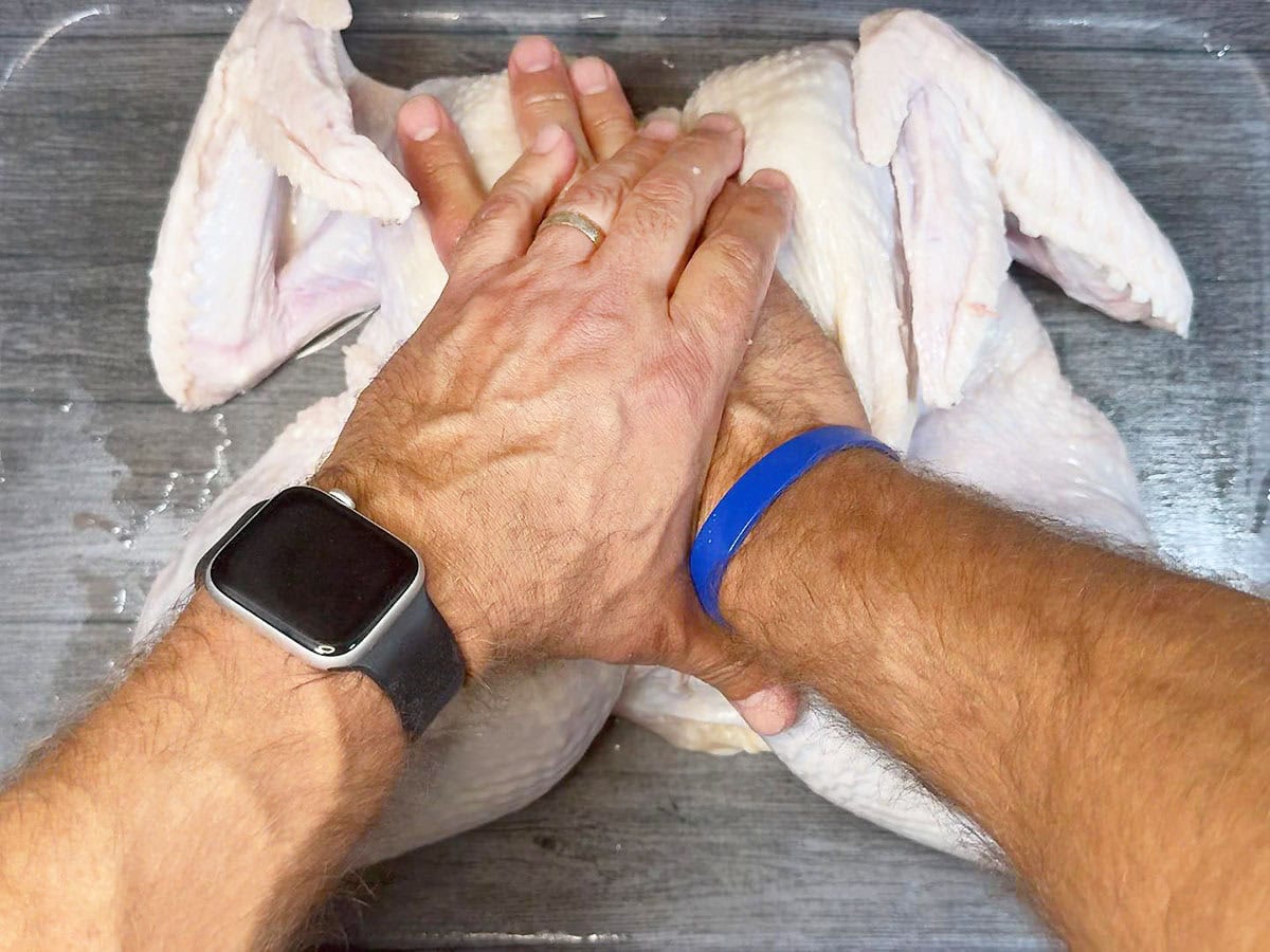 Hands pressing onto the breastbone of a spatchcocked turkey to flatten it.