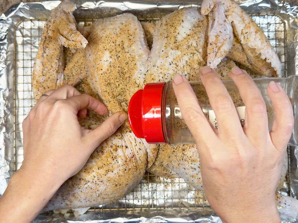Sprinkling all-purpose seasoning on the skin of an uncooked spatchcocked turkey.