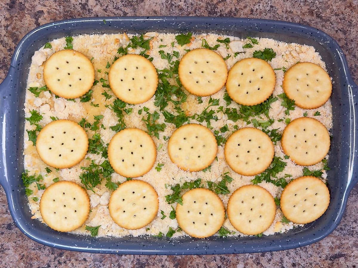 Unbaked scalloped oysters in a casserole dish topped with Ritz crackers, Parmesan cheese, and parsley.