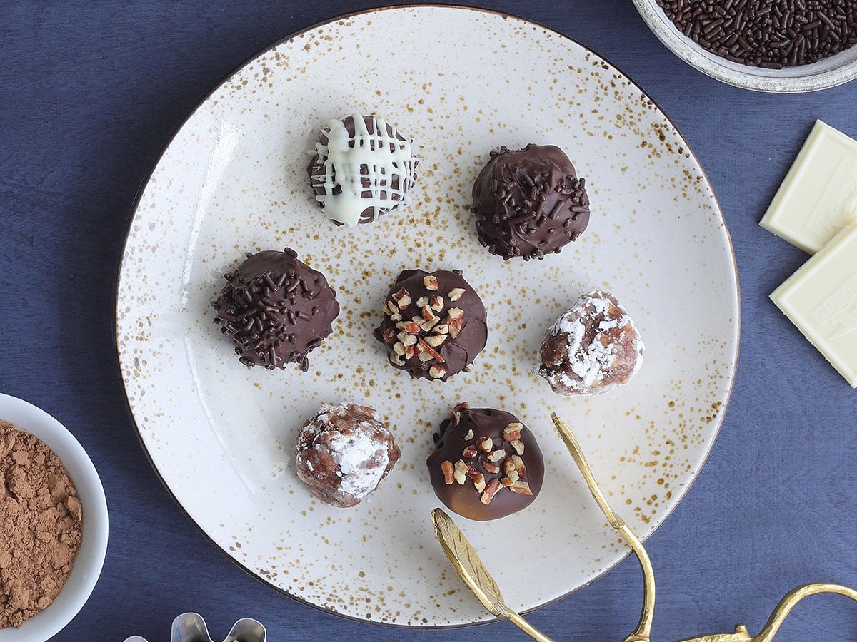 Seven decorated bourbon balls on a decorative plate with a set of tongs for serving.
