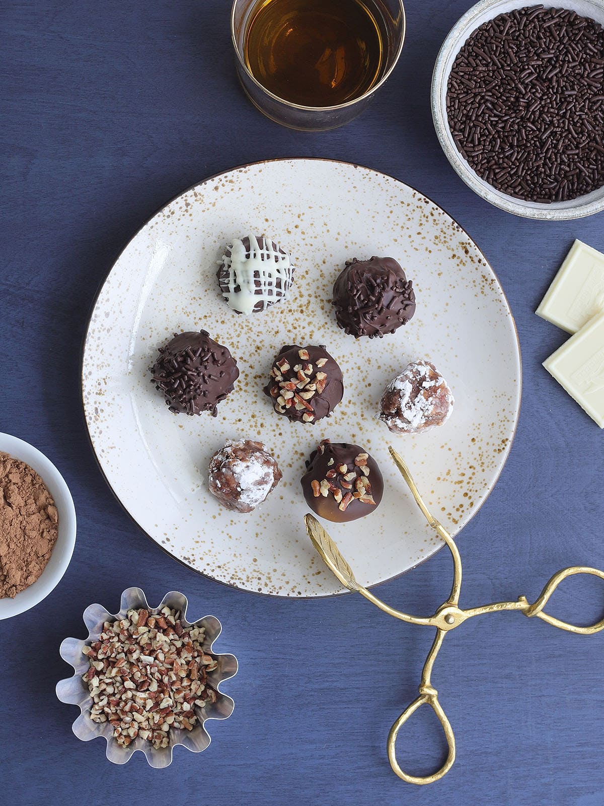 Seven decorated bourbon balls on a decorative plate with a set of tongs for serving.
