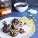 Seven decorated bourbon balls on a decorative plate with a set of tongs for serving.