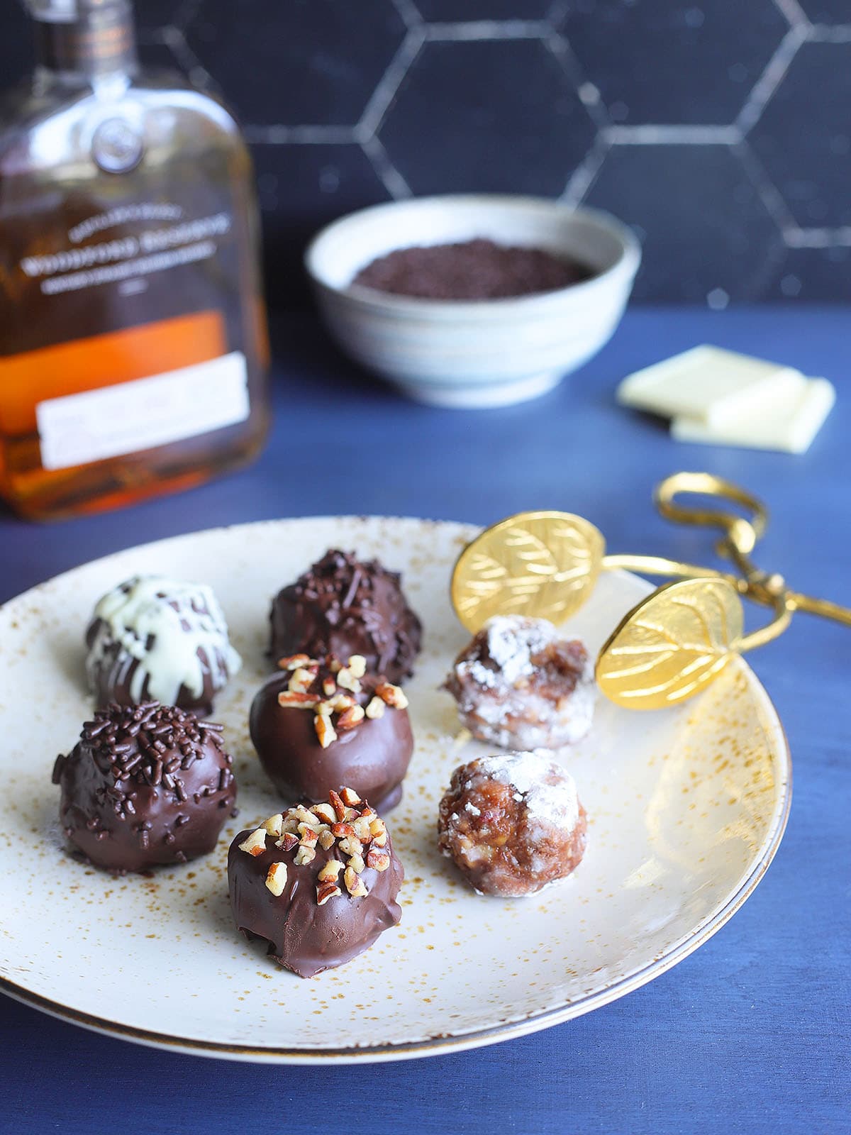 Seven decorated bourbon balls on a decorative plate with a set of tongs for serving.