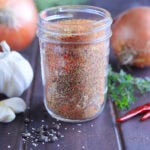Homemade Cajun seasoning in a glass Mason jar with onions, garlic and seasonings in the background.