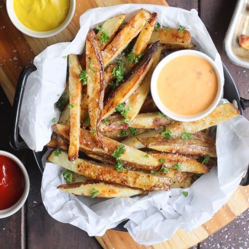 Oven baked French fries in a parchment paper lined dish garnished with salt and chopped fresh parsley.