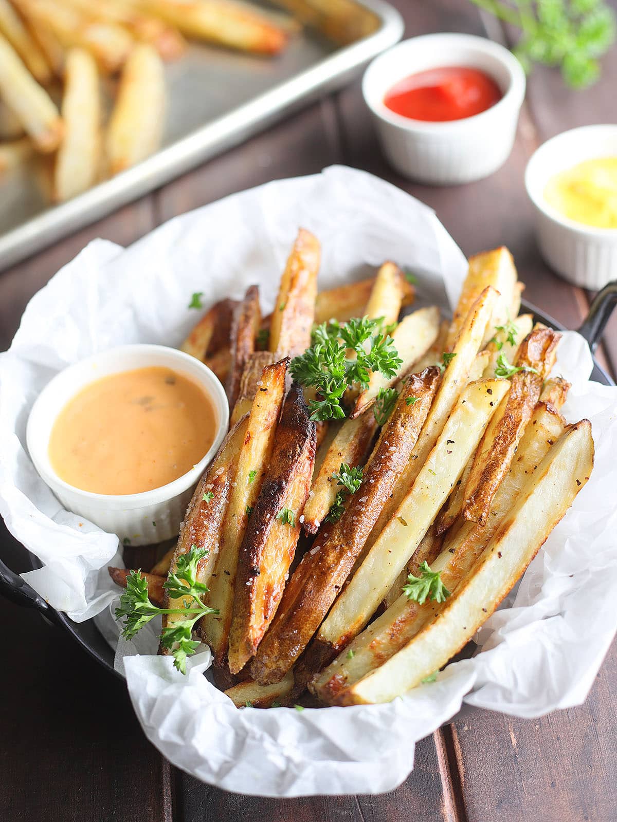 Basket of oven baked French fries garnished with salt and chopped fresh parsley with dipping sauces to the side.