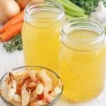 Two mason jars of shrimp stock with a small dish of shrimp shells and stock ingredients in the background.