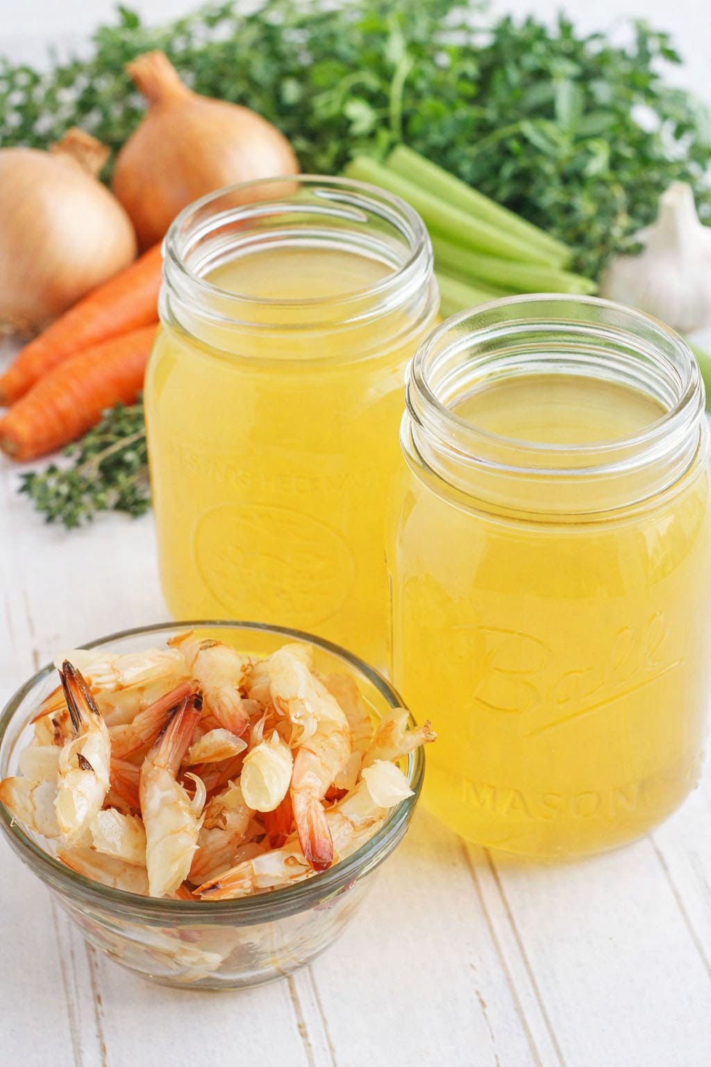 Two mason jars of shrimp stock with a small dish of shrimp shells and stock ingredients in the background.