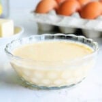 Dish of blender hollandaise sauce with a dozen eggs, stick of butter, lemon, and fresh parsley in the background.