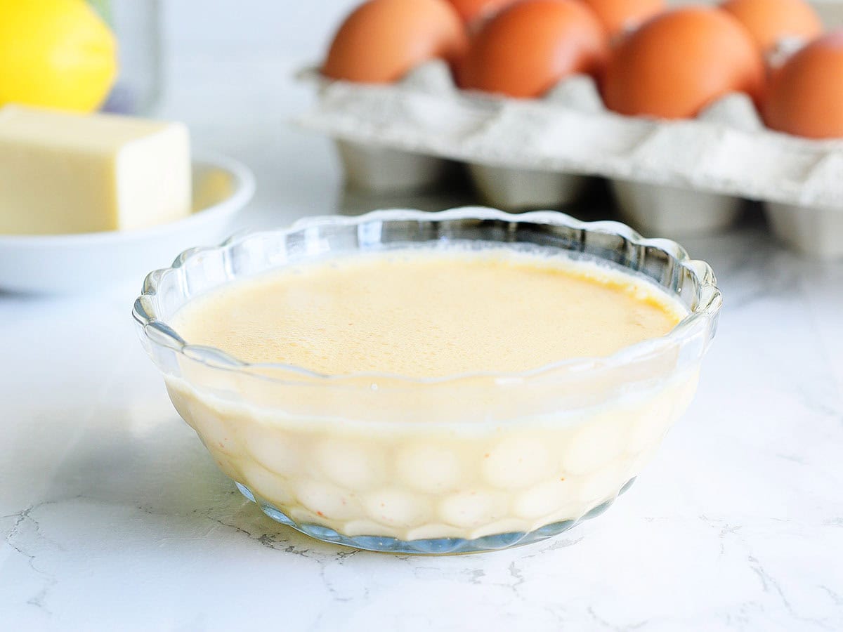 Dish of blender hollandaise sauce with a dozen eggs, stick of butter, lemon, and fresh parsley in the background.