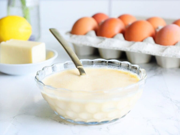 Dish of blender hollandaise sauce with a spoon. A dozen eggs, butter, lemon and parsley in the background.