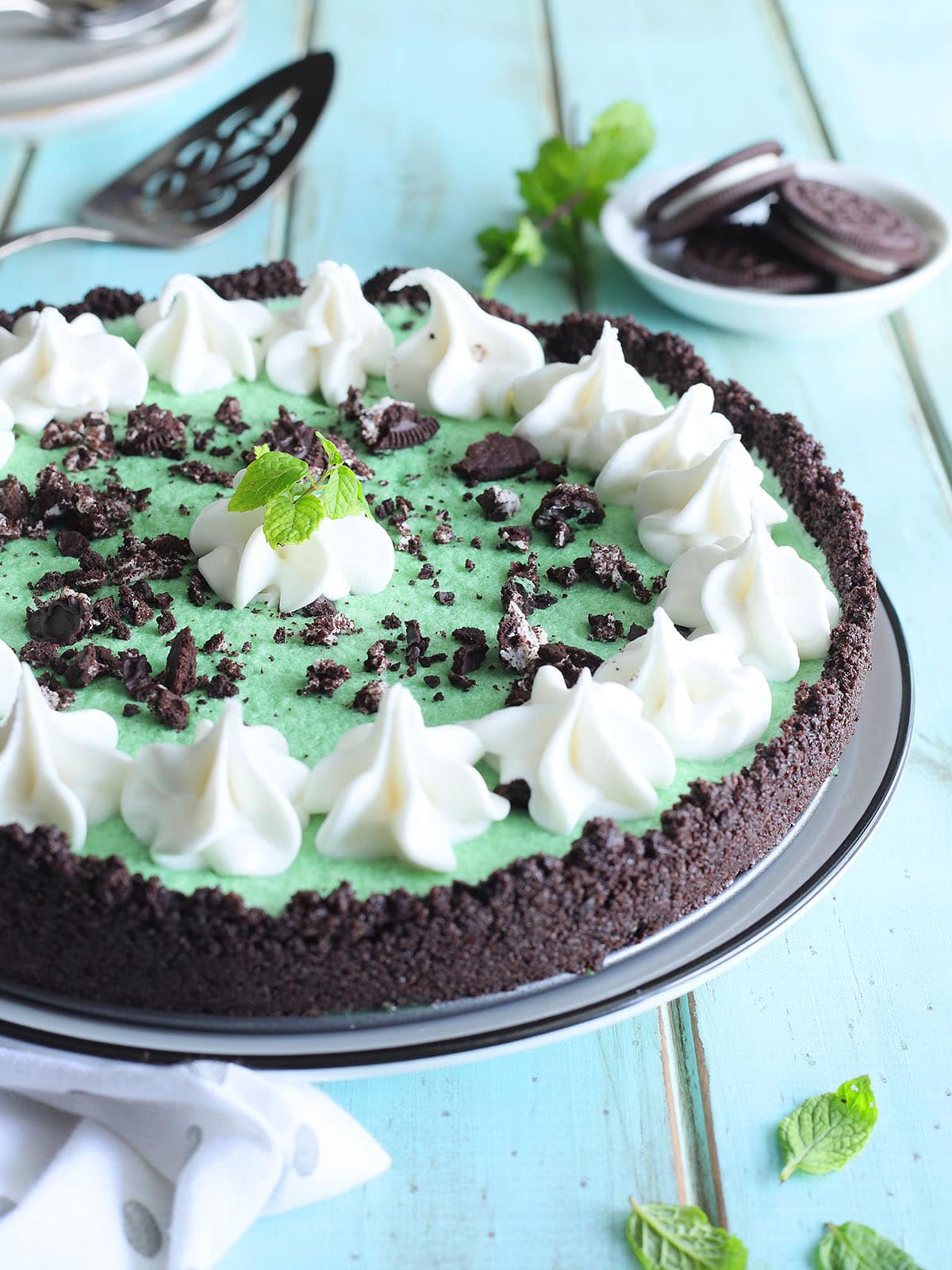 Grasshopper pie garnished with piped whipped cream, crushed cookies, and a mint sprig. A small dish of chocolate sandwich cookies and additional fresh mint are in the background.
