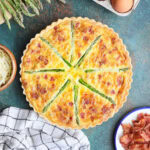 Whole asparagus quiche with eggs, fresh asparagus, crumbled bacon, and shredded cheese to the side.