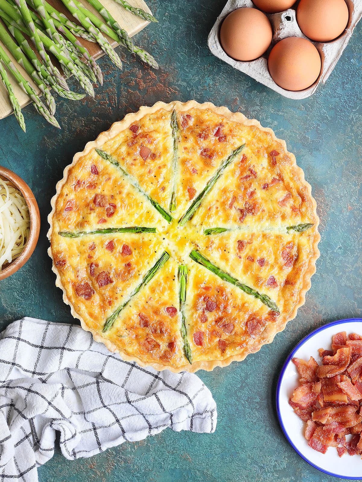 Whole asparagus quiche with eggs, fresh asparagus, crumbled bacon, and shredded cheese to the side.