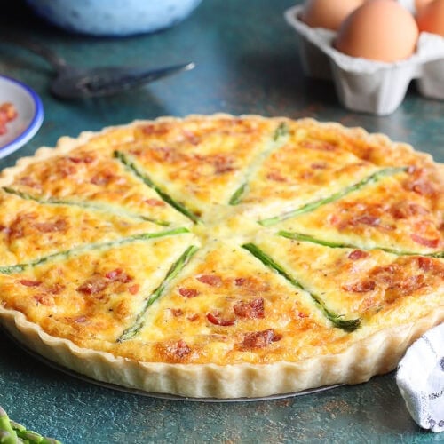 Asparagus quiche with eggs, crumbled bacon, and fresh asparagus in the background.