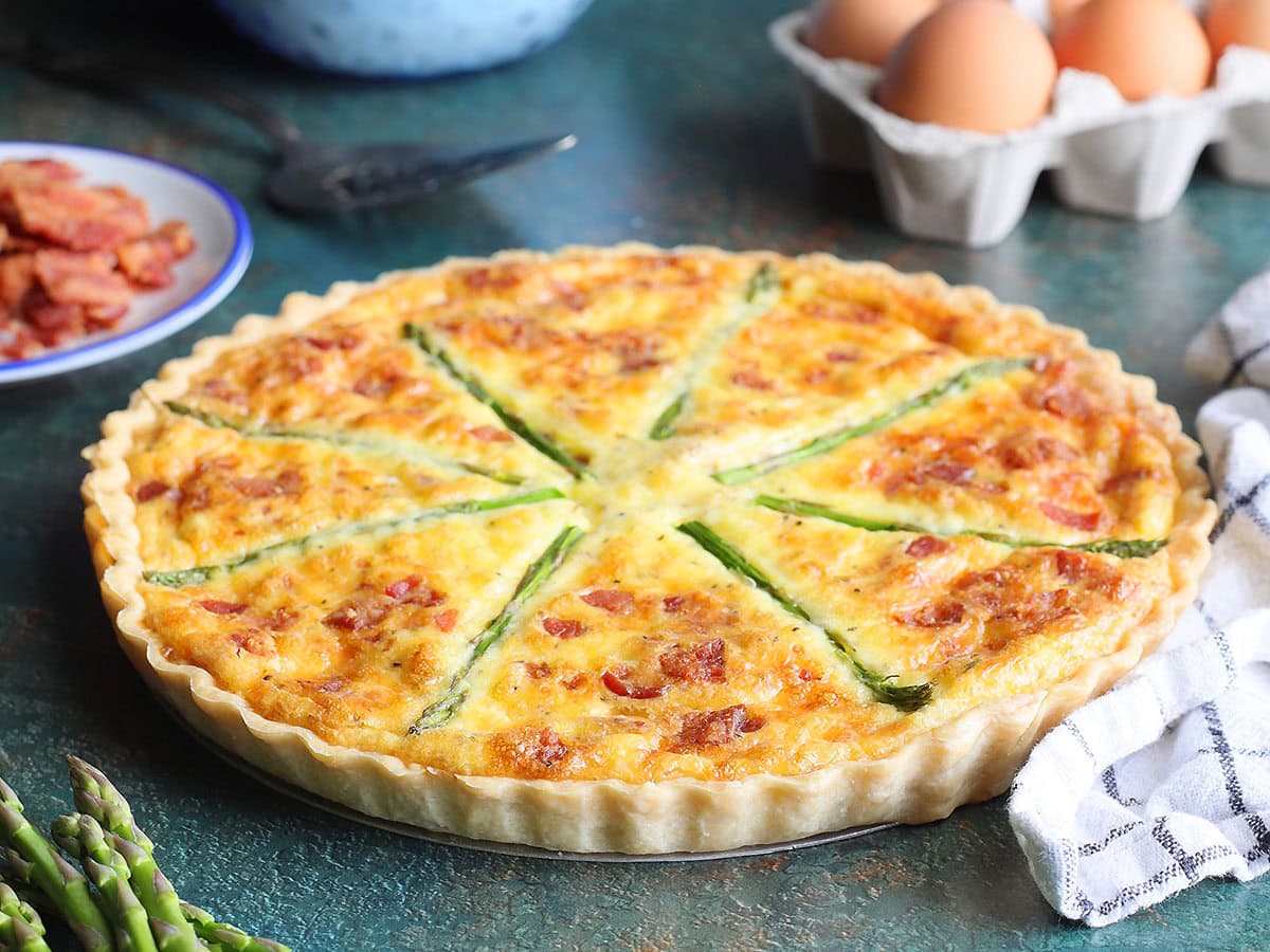 Asparagus quiche with eggs, crumbled bacon, and fresh asparagus in the background.