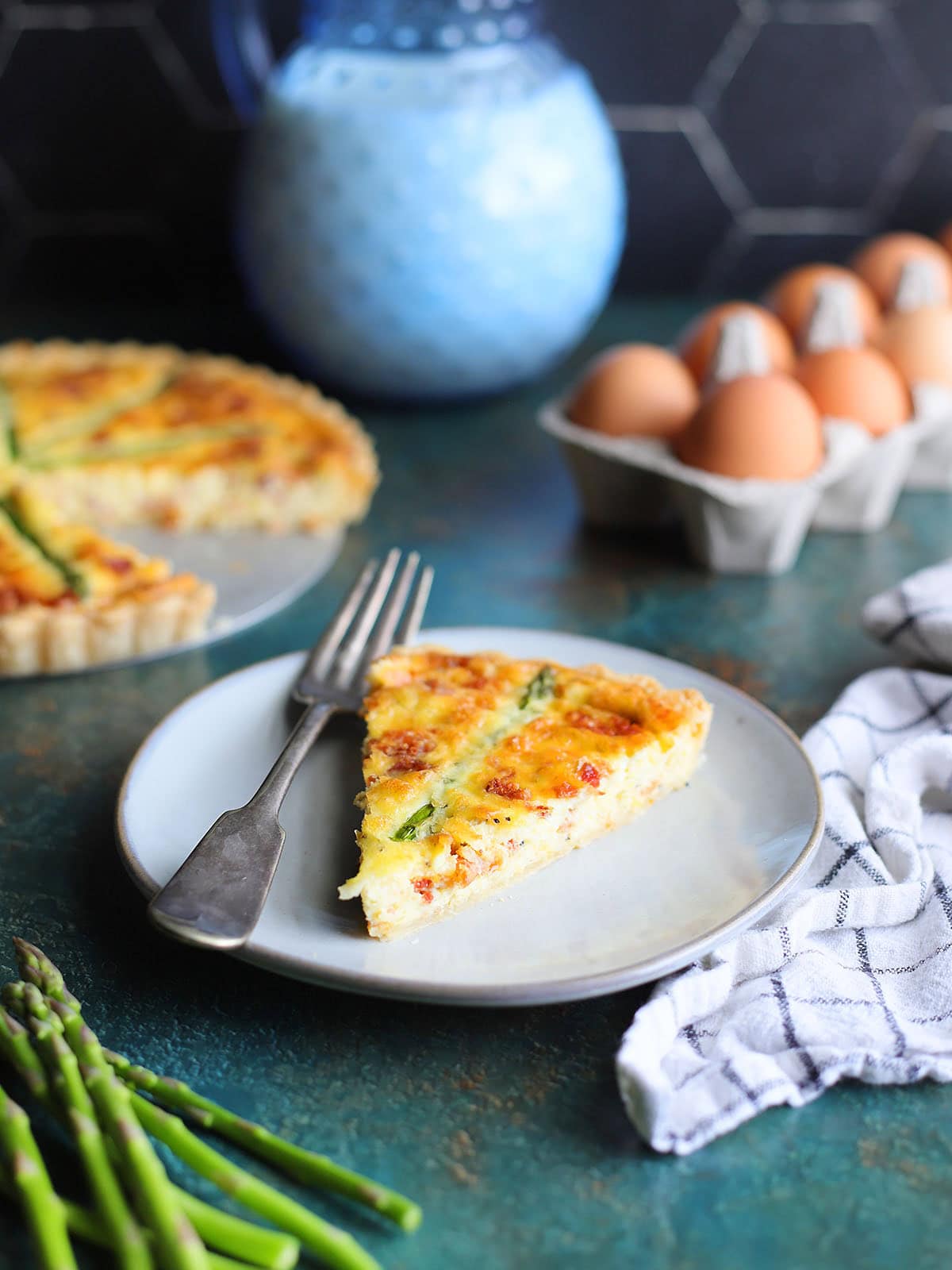 Slice of asparagus quiche on a plate with a fork with a dozen eggs, a pitcher of milk, and the sliced quiche in the background.
