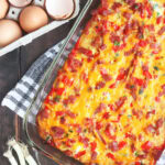 Baked breakfast ham casserole with tater tots on a wooden tabletop with a dozen eggs and three green onions on the side.